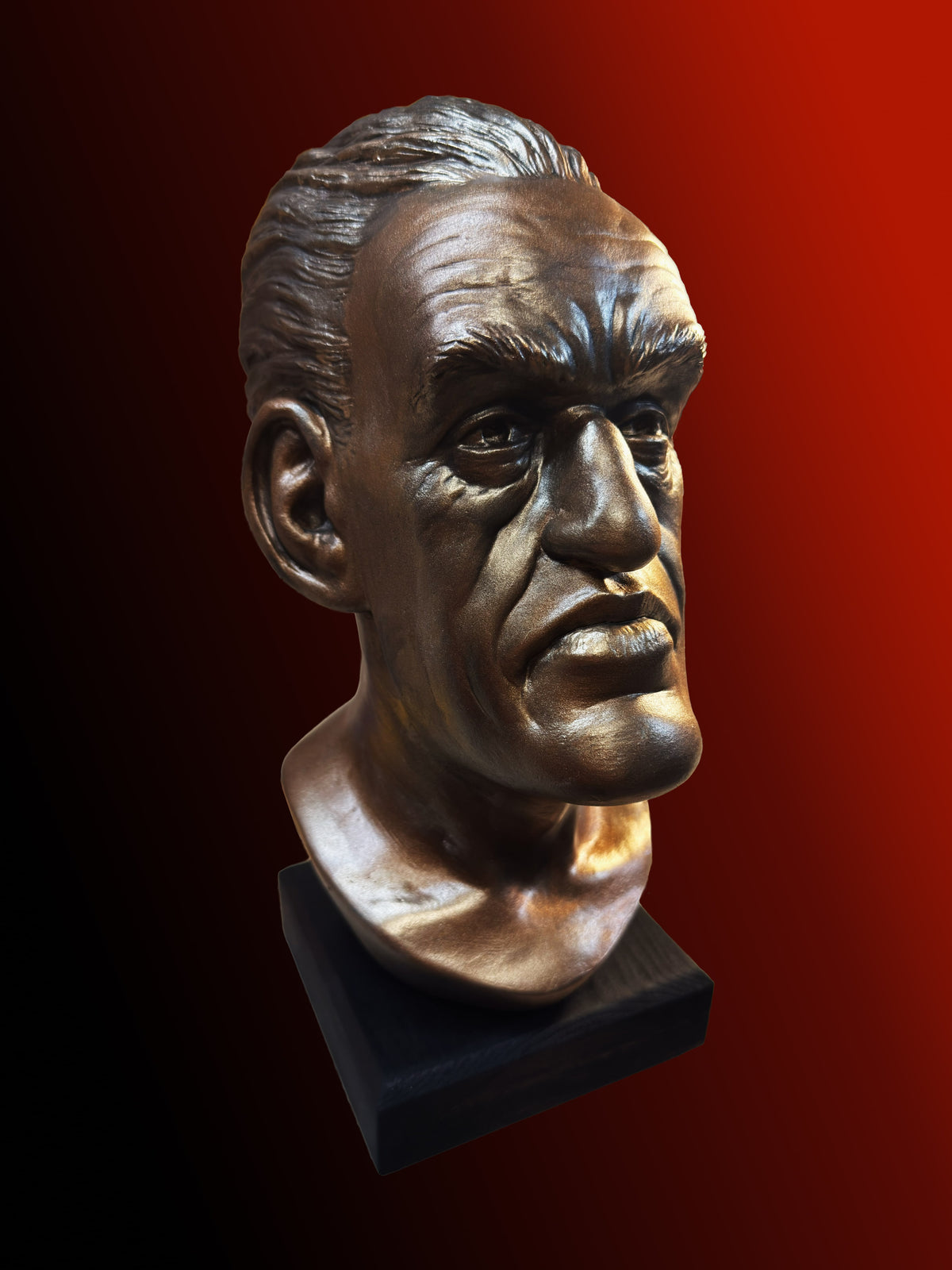 A bronze-finished bust sculpture of a man's head mounted on a black rectangular base, shown against a dark red to black gradient background.