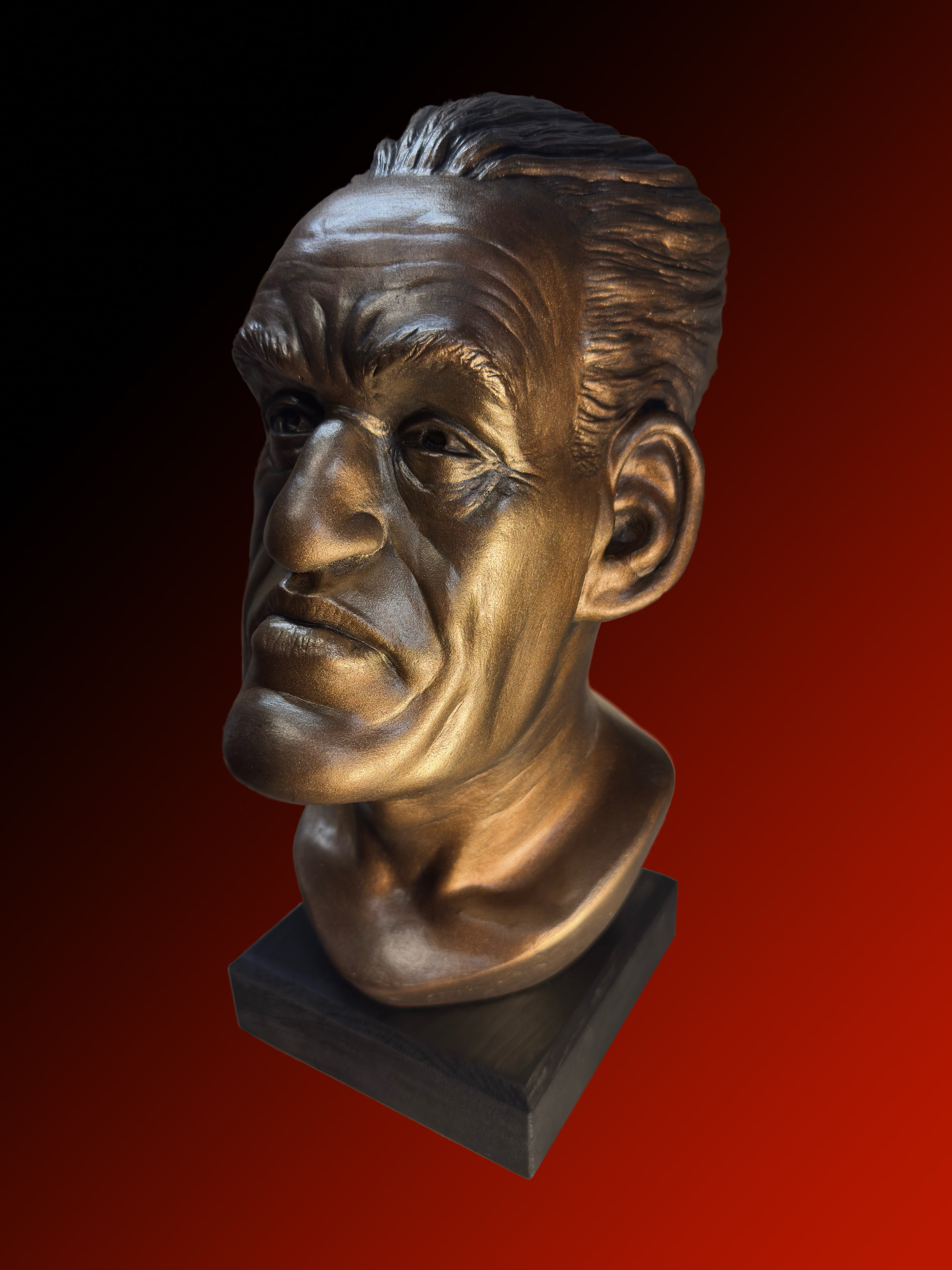 A metallic bronze bust sculpture of a man's head mounted on a dark rectangular base, shown against a red-to-black gradient background.